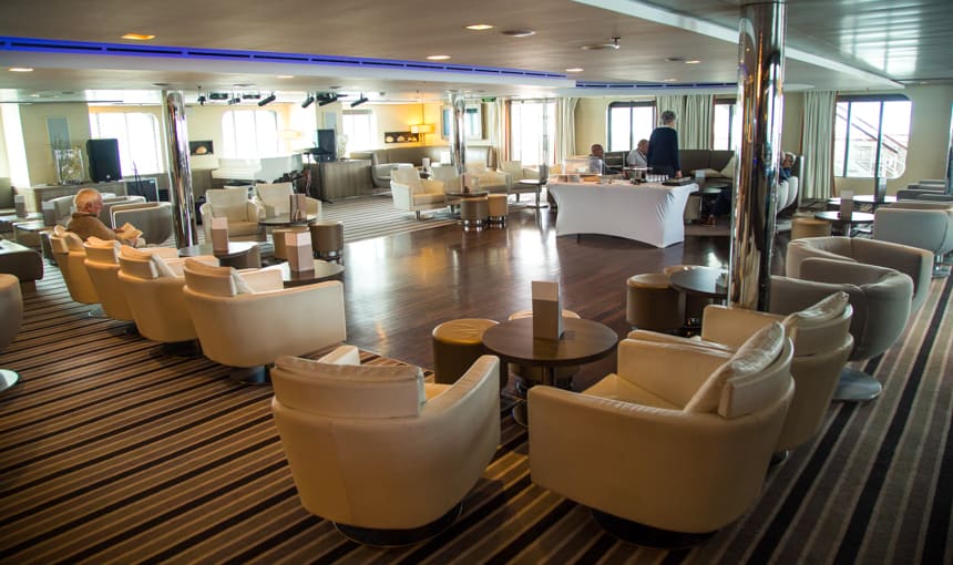 A lounge seating area aboard L'Austral and her ponant luxury sister ships. surrounded with large glass windows cream, beige and brown leather arm chairs are set facing the windows with ottomans and coffee tables on top of striped carpet and wooden square dance floor.