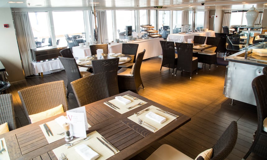 One of the restaurants and dining room options aboard L'Austral and her ponant luxury sister ships. surrounded with large glass windows brown tables and chairs are set around a centered steel buffet area.