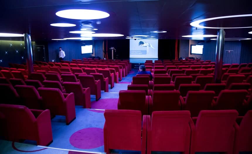 the theater inside L'Austral and its sister ships with rows of red comfy movie theater seats lined in a circular fashion, facing the front stage, the lights are dimmed and a screen at the frotn is showing an educational video. circular lights on the ceiling match the large circular dot pattern on the carpet.