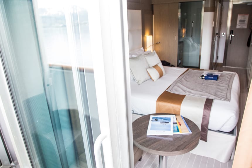 Standing outside on a private cabin balcony looking in is a cabin aboard L'Austral luxury ship, an accent chair and coffee table, queen bed with white linen.