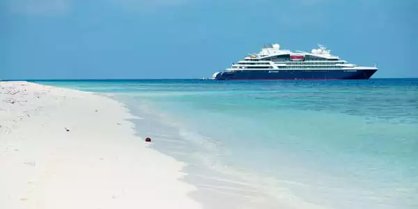 Luxury Caribbean cruise ship with blue & white exterior sits offshore of a private island white-sand beach.