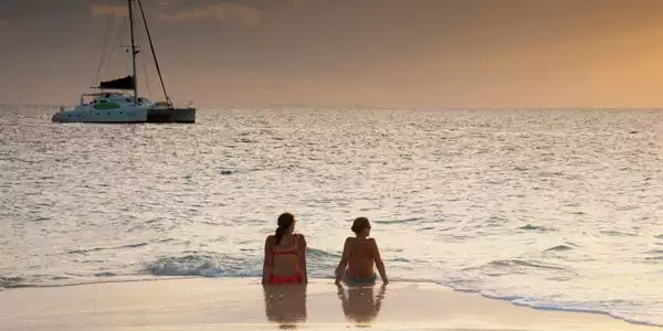 2 women sit at water's edge on a sandy beach at sunset with their private Caribbean catamaran charter yacht in the distance.