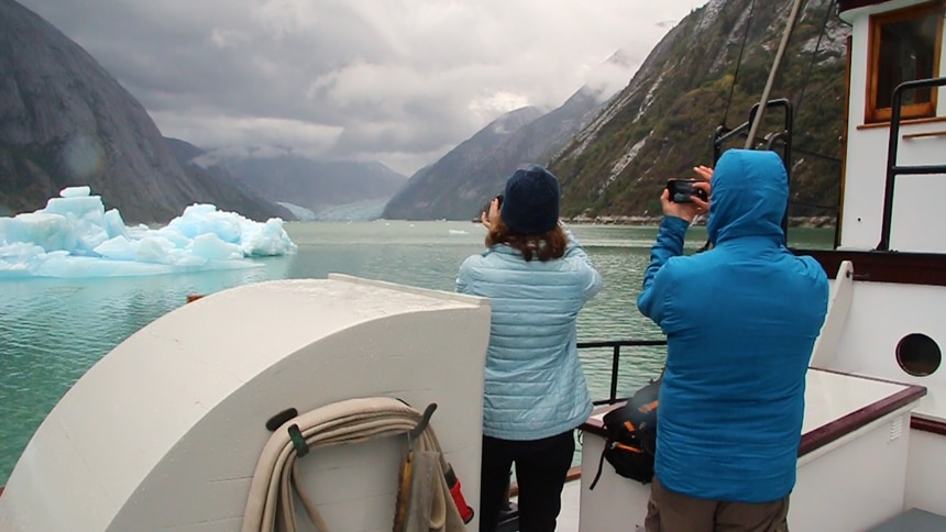 two cruise guests stand on the front desk of an Alaska small ship, with their cameras raised, they photograph the distant icy glacier.