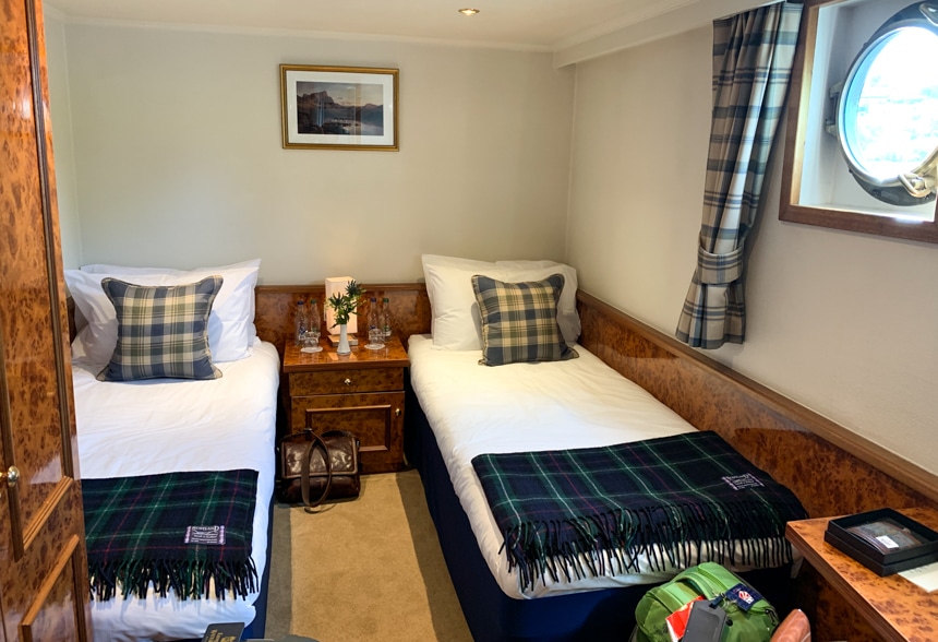 A cabin aboard Spirit of Scotland Barge, two double beds share a wooden end table, plaid pillows and blankets red on the bed, along with plaid window curtains for the porthole window.