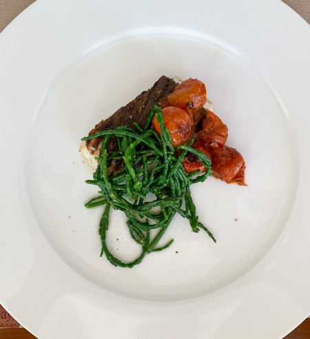 A dinner plate served aboard the Classic Scotland Barge Cruise, a piece of grilled fish with roasted red tomatoes and green vegetables sit on a white plate.