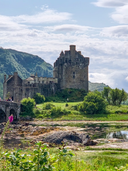 On top of a small, lush green island connected by a stone bridge sits a Scottish stone castle, seen aboard the Classic Scotland Barge Cruise