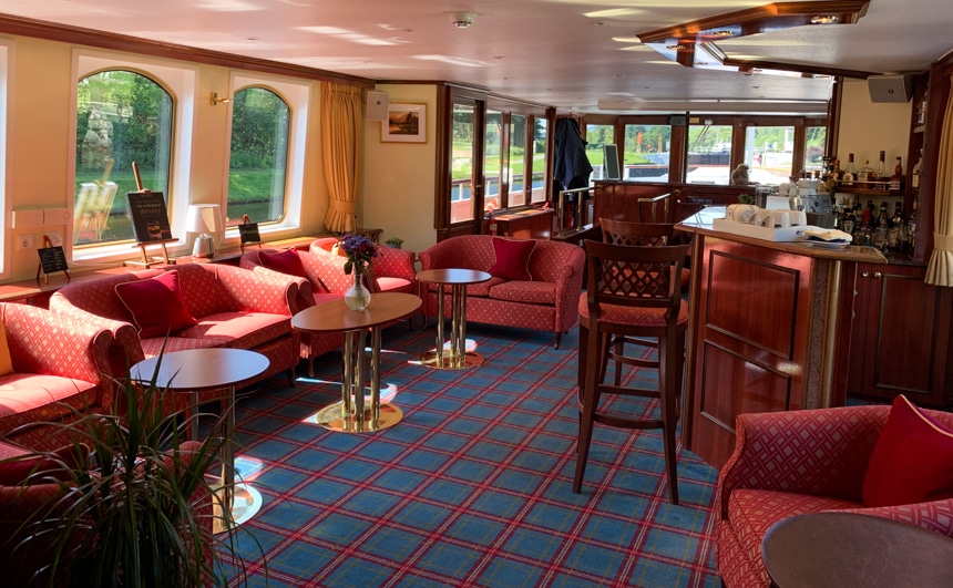 Inside the lounge aboard Spirit of Scotland Barge, plaid carpets sit underneath a set of red love seats and arm chairs, with wood and gold coffee tables, surrounded by glass windows.