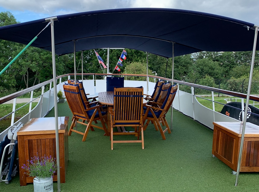 Covered dining terrace on the rooftop deck of Spirit of Scotland barge with short green turf carpeting, and a wooden table and chairs.