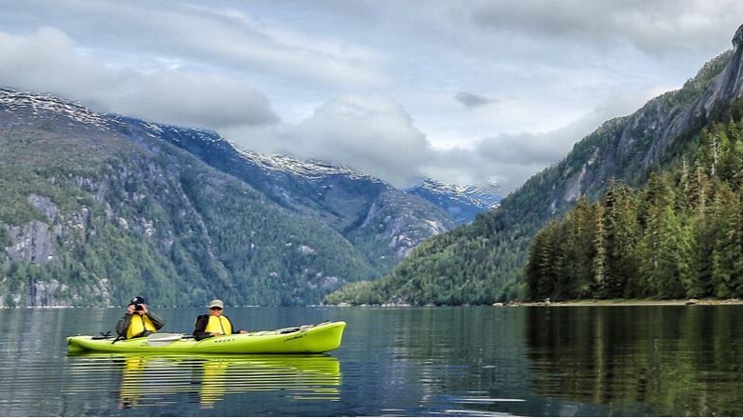 Tandem kayakers in a bright green kayak practice their Alaska cruise photography in calm waters with mountains in the background on a partly sunny day.