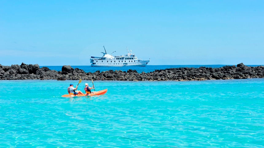 In the Galapagos Islands two kayakers paddle their kayak in teal ocean water, floating in the distance is small ship La Pinta.