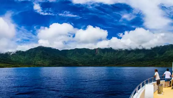 Man stands on bow of small ship looking at deep blue sea & verdant green hillside on a partly cloudy but bright day during a small ship cruise in the Pacific Islands.