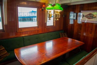The main salon aboard Alaska small ship Catalyst. A long wood table with green bench seating and windows to the outside,