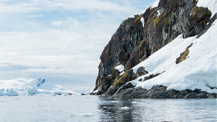 Zodiacs with Antarctica travelers sit in the distance, beside a small rocky mountain with snow.