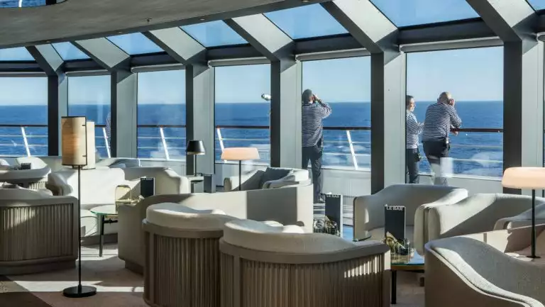 Observation Lounge on Le Commandant Charcot lng ship with beige chairs by floor-to-ceiling windows showing guests on deck in the sun.