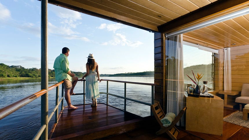 Woman & man stand on bow of private balcony on wooden Amazon riverboat as it cruises.
