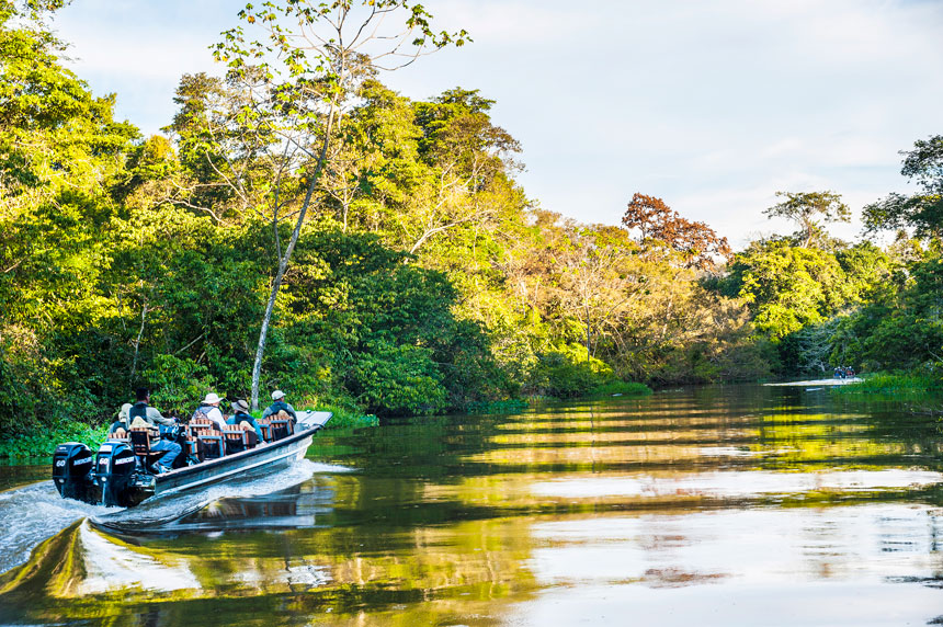 Amazon travelers & guide ride in an aluminum skiff boat along glassy water lined by jungle.