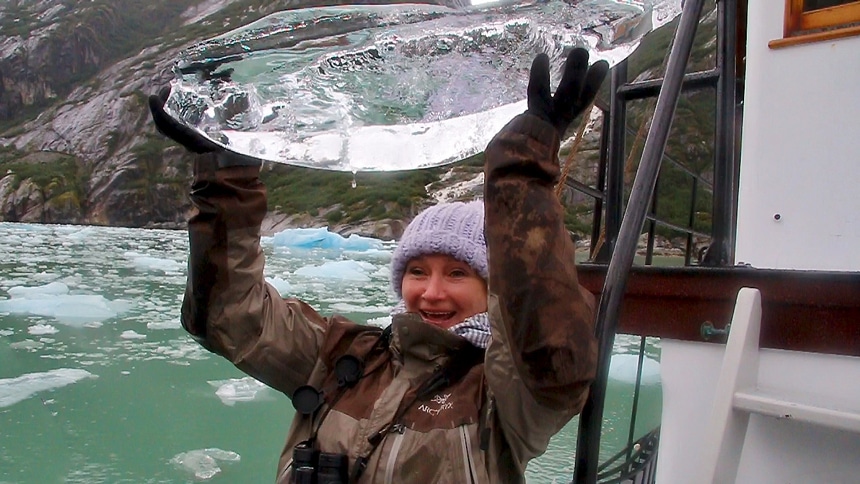 Aboard Catalyst in Alaska's Endicot Arm a female cruise guest lifts a large crystal clear piece of iceberg above her head 