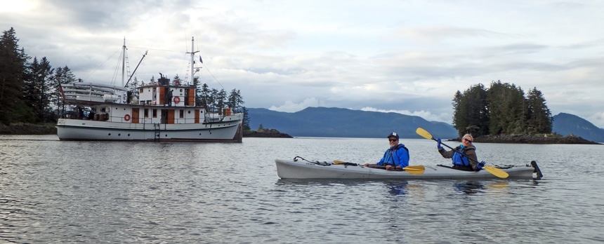two travelers paddle a gray double kayak in front of the small ship Catalyst on their slow travel cruise in  Alaska