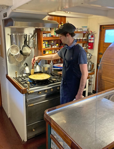 An open kitchen aboard Alaska small ship catalyst, the chef wears a blue apron and holds a spatula in a pan filled with food. 