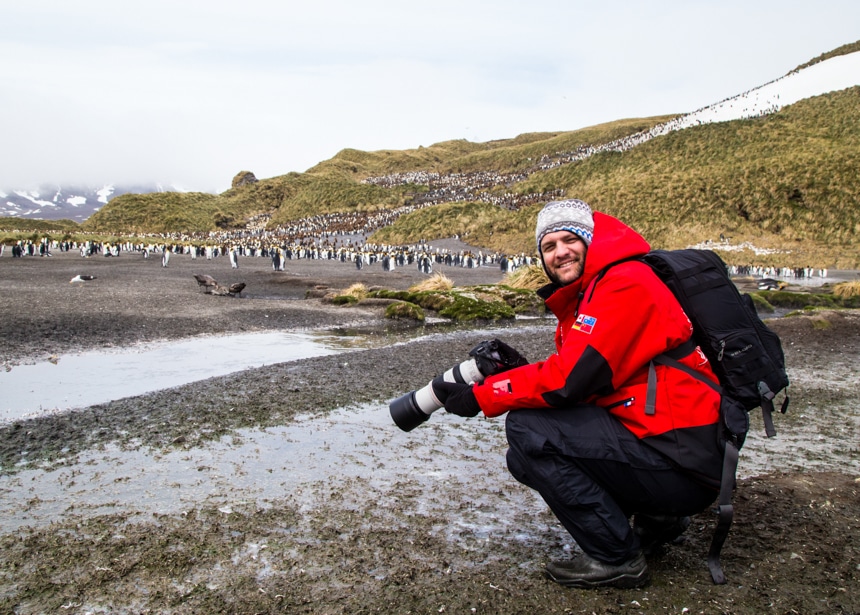 a man kneeling down and smiling as he takes photos of the large, scattered king penguin colony in antarctica