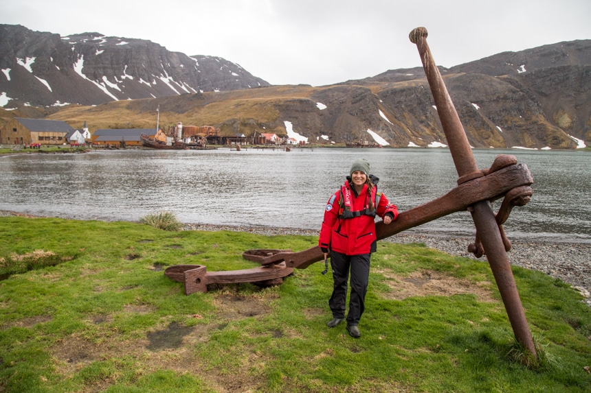a woman standing in front of a large, rusted ship anchor that sits on the grassy shore in grytviken south georgia, antarctica