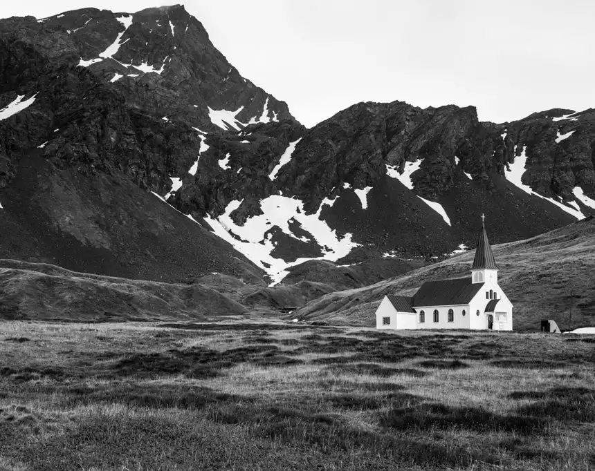 a black and white photo of church that sits among grassland below the tall mountains in grytviken, south georgia