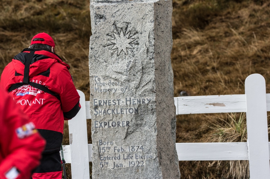 a person standing in front of shackelton's grave stone that stands in front of a white fence in grytviken, south georgia