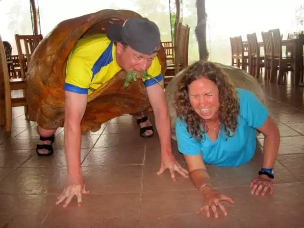 Two laughing people on the ground wearing large tortoise shells. 