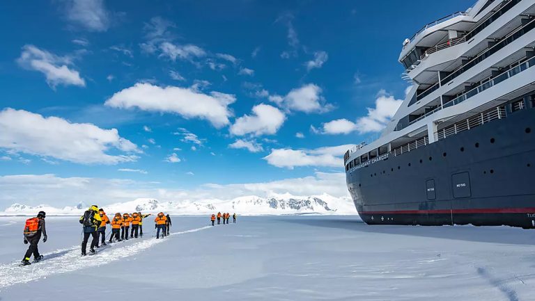 Le Commandant Charcot is parked in an ice sheet in Bellingshausen sea, travelers walk through snow on the ice around it,