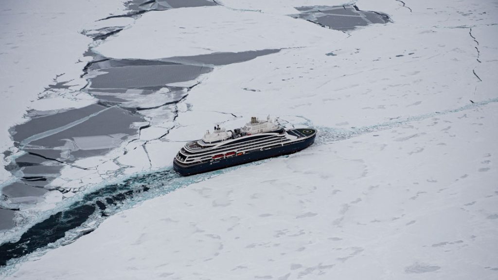Aerial view of dark blue expedition ship cruising ice-laden waters on a stormy day.