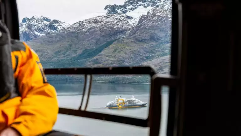 Chile cruise guest in yellow jacket sits in helicopter looking at Patagonia's peaks & Ultramarine expedition ship down below.