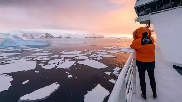 Woman in orange parka with fur-lined hood stands on deck of white ship photographing sea ice at sunset in the Bellingshausen Sea.