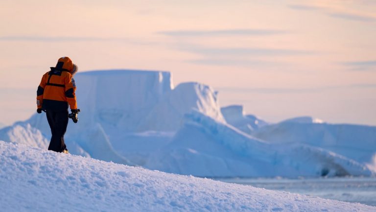 In the Ross Sea a traveler in red fur lined hooded parka walks through a snowy icy Antarctic landscape during a pastel sunset,