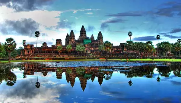 Beautiful temples reflect off of glassy water, surrounded by tall palm trees under a blue sky, on a Southeast Asia vacation.