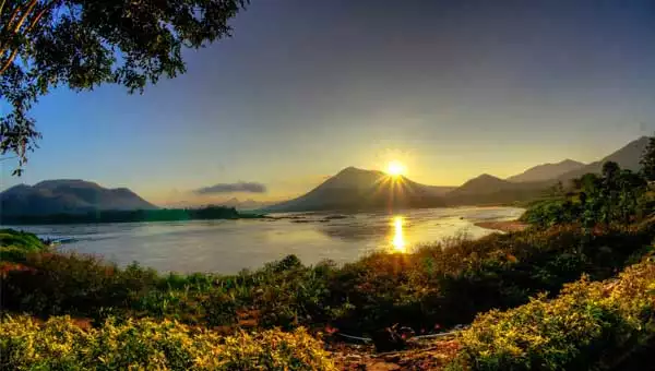 Sun rises above volcanic mountains with bright green bushes in the foreground, seen during a Mekong River cruise.