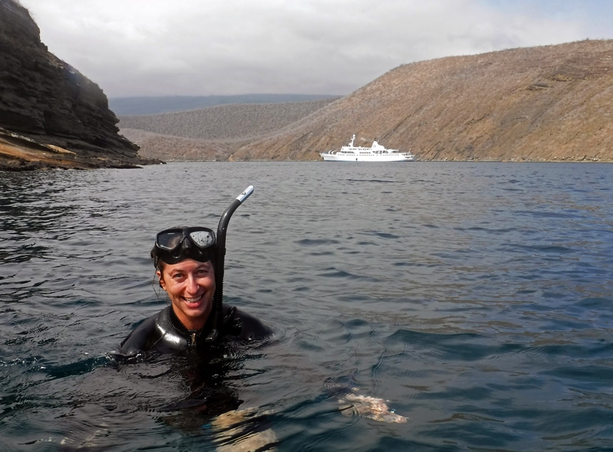 A female snorkeler floats at the surface of the dark ocean wearing a black wetsuit, mask and snorkel, behind her is a small Galapagos ship.