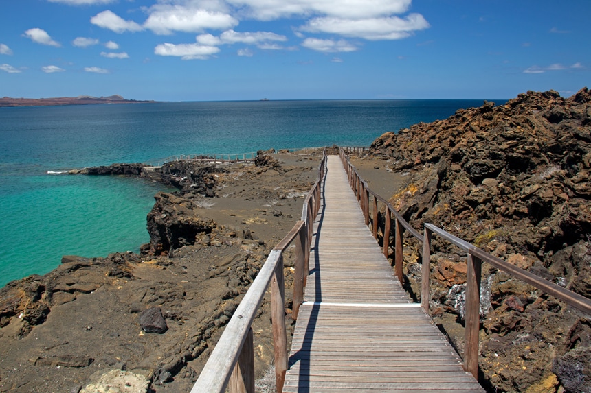 Over jagged lava rock of the Galapagos a wooden pathway with hand rails leads down to the teal ocean waters edge.