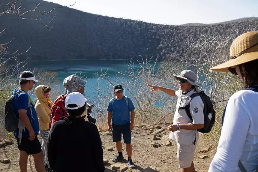 A Galapagos naturalist guide wears a tan uniform with wide brimmed hat speaks to his group and points to the landscape around them.