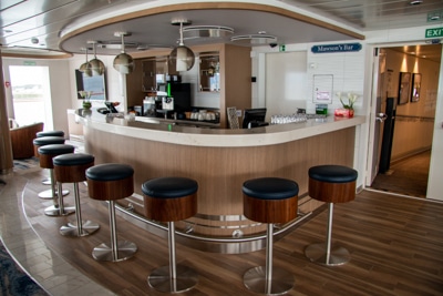 A set of bar height stools sit around the bar aboard Greg Mortimer Antarctic ship, circular light fixtured hang from the ceiling.