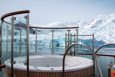 A jacuzzi surrounded half way with glass sits on the top deck with views of the snowy Antarctica landscape the ship cruises by.