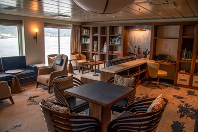 Library and lounge aboard Greg Mortimer ship, computer desks, artwork, shelves filled with books accent chairs and seating areas.