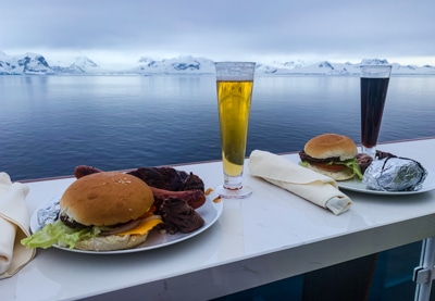 Dinner with a view. Plated food and tall drink glasses are set on an outdoor table facing the ocean towards snow capped mountains of Antarctica