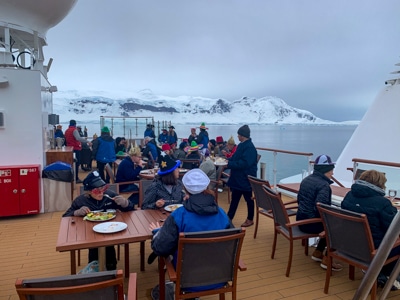 On the top deck of Greg Mortimer ship, guests enjoy their meal outside at tables with views of Antarctica's snowy mountain ranges.