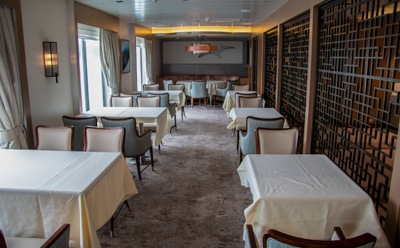 The empty dining room aboard Greg Mortimer Antarctic Ship, tables covered with table cloths and upholstered accent chairs set in rows.