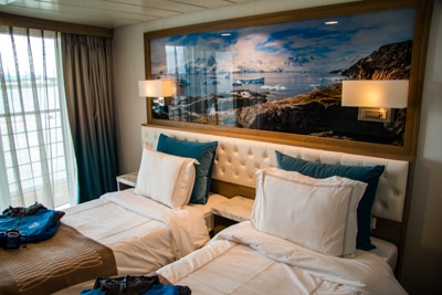 Stateroom aboard Greg Mortimer ship, two double beds, sliding glass balcony doors, and a large Antarctica landscape framed photo overhead.