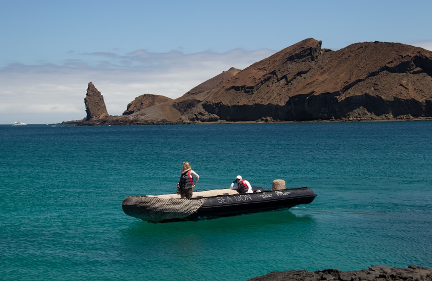 Pinnacle Rock, a volcanic plug on Bartolome Island, rises from the teal ocean as a black inflatable skiff cruises around in front of it.