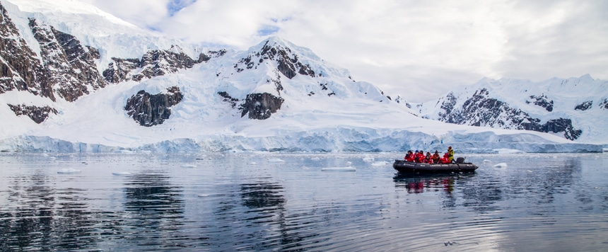 Un grupo con parkas de invierno rojas se sienta a bordo de un crucero en bote inflable negro a lo largo de la costa nevada de la península antártica.