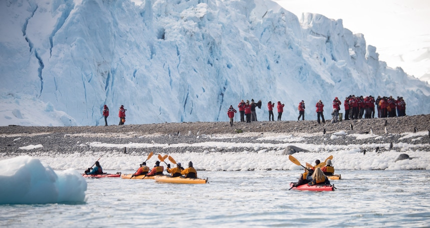 A group of cruise passengers wearing red parkas walk on shore while others paddle in double kayak as activity options offered during Antarctica cruses.