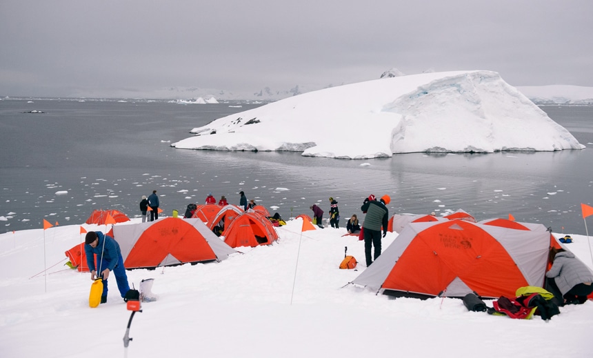 En un crucero de un día nublado gris, los huéspedes instalan sus tiendas de campaña naranjas en la costa de la Antártida para una actividad de campamento polar