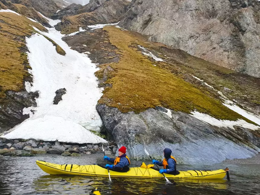 Against the shore line of an Arctic island in Svalbard two guests wearing winter gear paddle a yellow double kayak through the ocean.